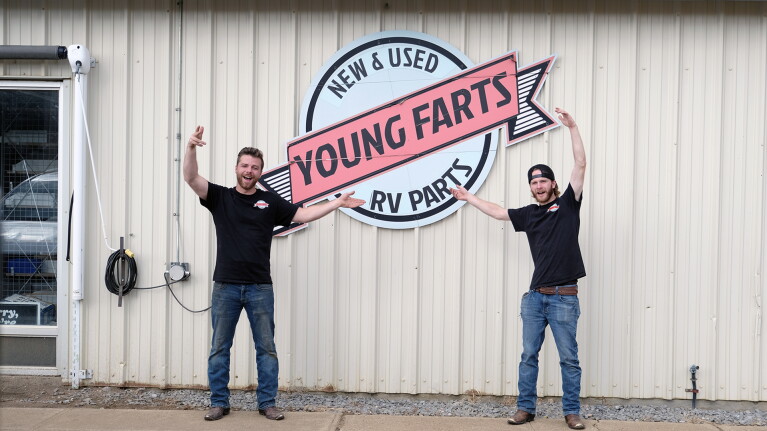 Two men stand outside a yard with the sign 'New & Used Young Farts RV Parts'