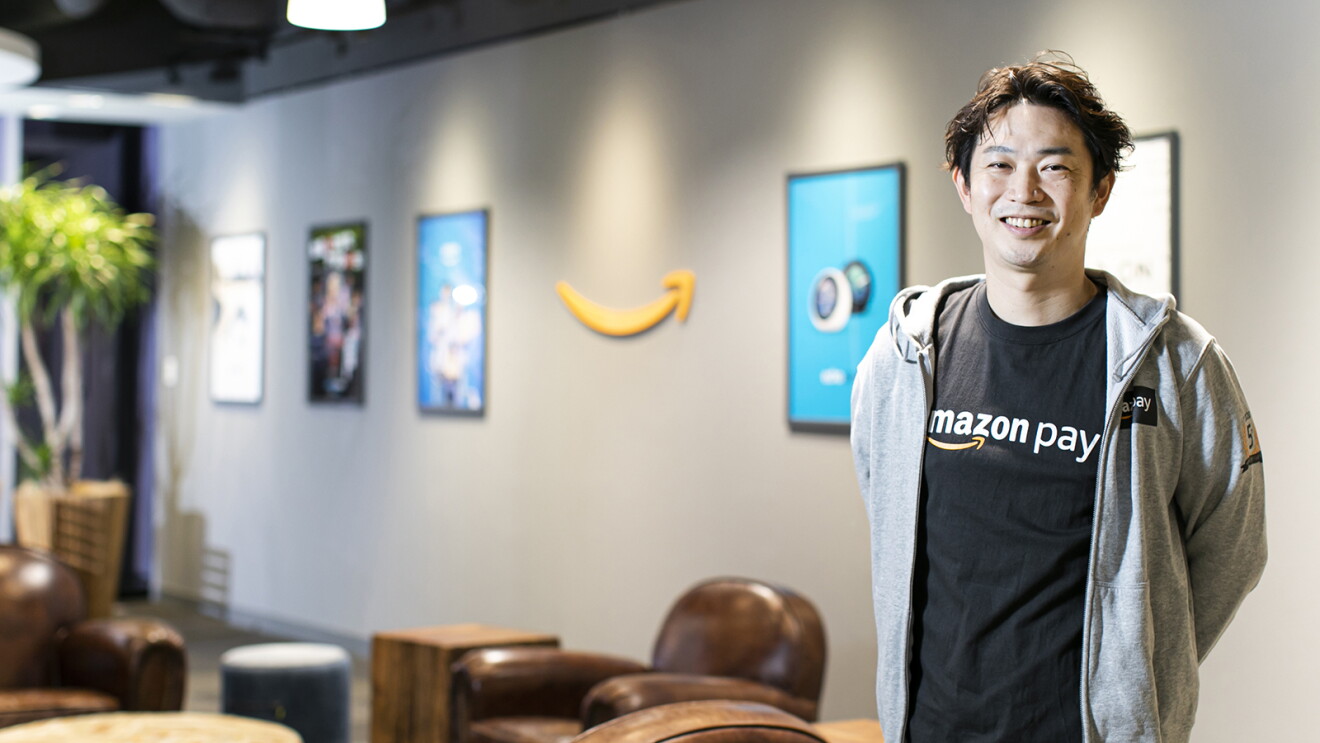 薄いグレーの壁の室内に立つ男性。グレーのパーカーに黒いシャツ。壁みは縦長のパネル入りポスター、黄色いAmazonロゴが設置されている。