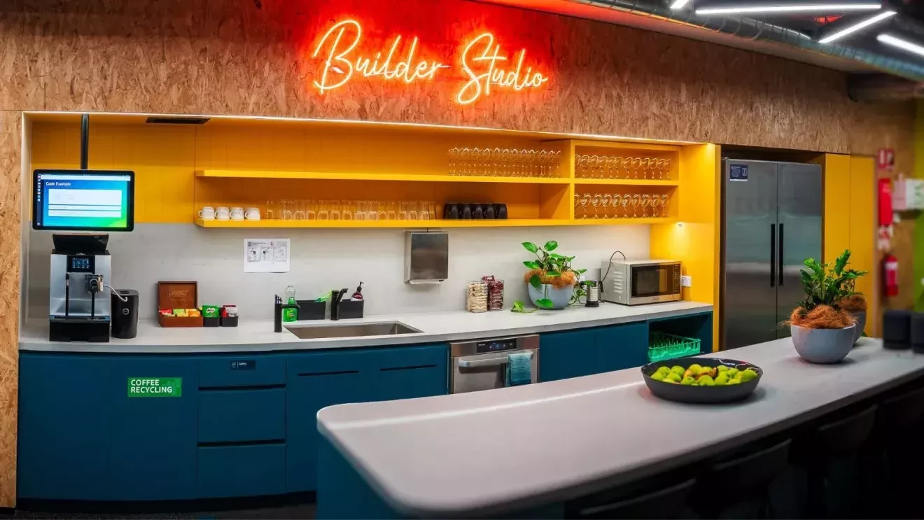 ビーチの雰囲気を演出している黄色と青のカウンター。そのカウンターの上に取り付けられている「Builder Studio」のサインが光っている。コーヒーメーカーや果物などがカウンターのテーブルの上に置かれている。