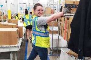 A woman wears a yellow safety vest and glasses as she puts am Amazon box on a shelf in a fulfillment center.