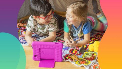 An image of two children laughing and interacting with an Amazon tablet. They are laying down on a colorful carpet and have toys sitting on the floor around them. 