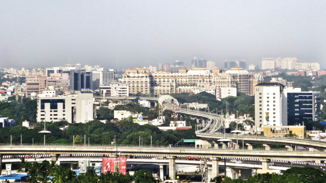 Aerial view of Chennai cityscape with multi-lane highway, metro rail line, and modern buildings surrounded by greenery