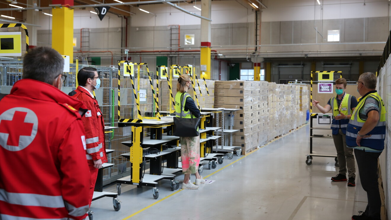 Links im Bild: ÖRK-Vertreter in den roten Jacken des österreichischen Roten Kreuzes. Im Bildhintergrund: Logistikmitarbeiter in Sicherheitswesten und mit Masken.