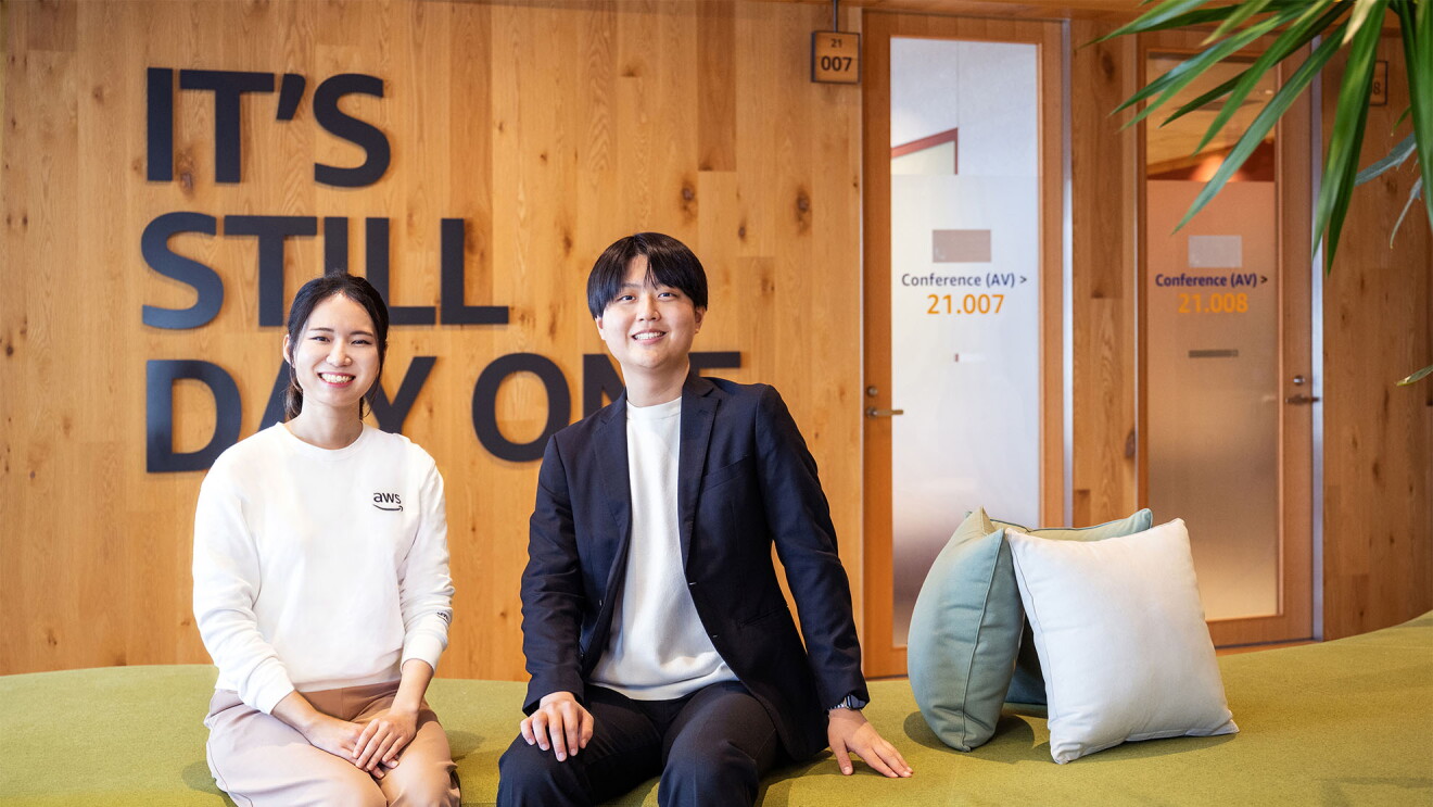 モスグリーンの大きなソファに2名が着席している。右側の人物はAWSロゴ入りの白いスウェットとピンクベージュのパンツを着用。左側の人物はネイビーのジャケットセットアップ。人物の右側に3つのクッション。背後の壁面は木製で、「IT'S STILL DAY ONE」の黒い文字で装飾されている。右奥には会議室へと続く2つのドアと観葉植物が見える。