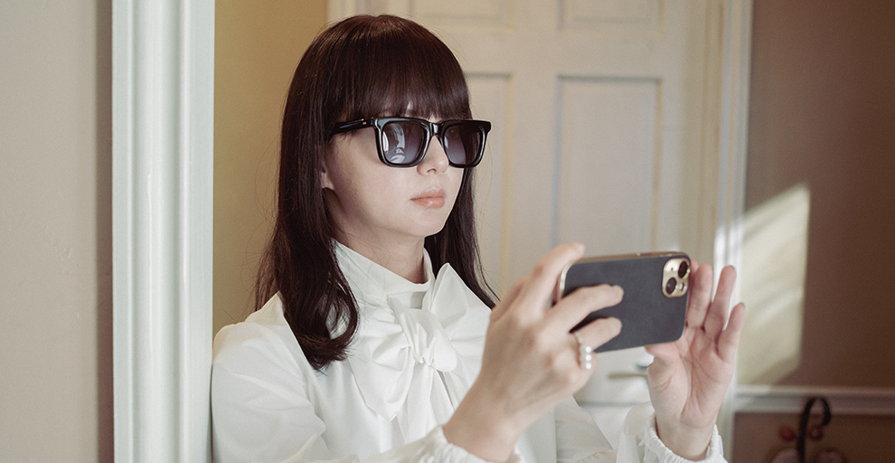 室内で大きなサングラスをかけた女性。スマホを横長に構えて何かを撮影しようとしている。