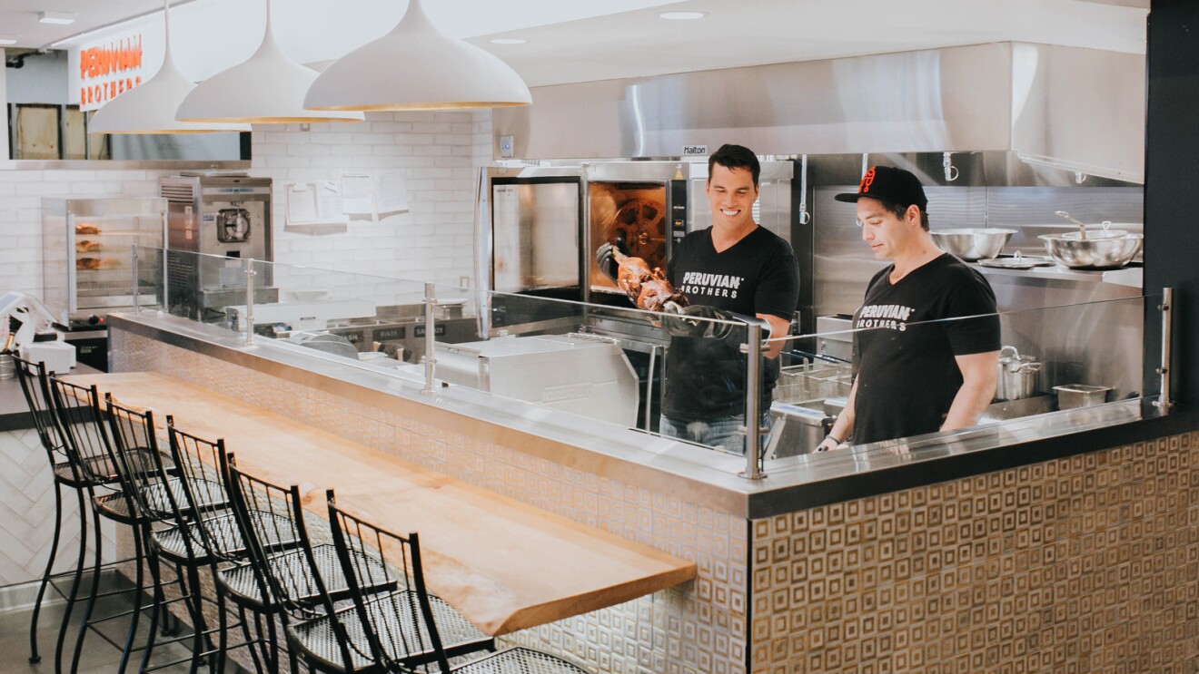 A photo of Mario and Giuseppe Lanzone working in The Peruvian Brothers catering kitchen next to a bar that seats six chairs.