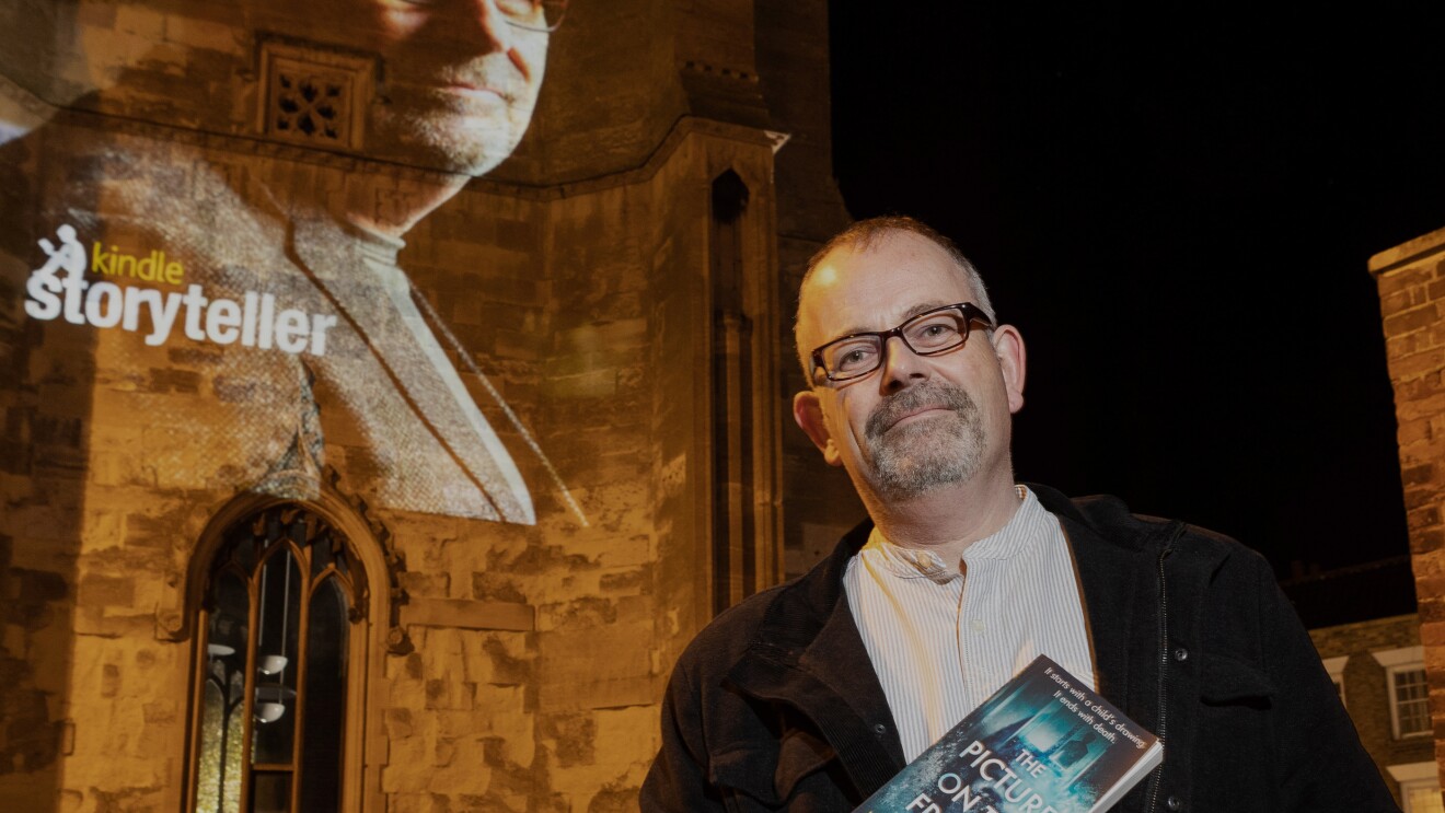 Ian W. Sainsbury holding his book in front of a Storyteller projection of his head shot congratulating him on his award.