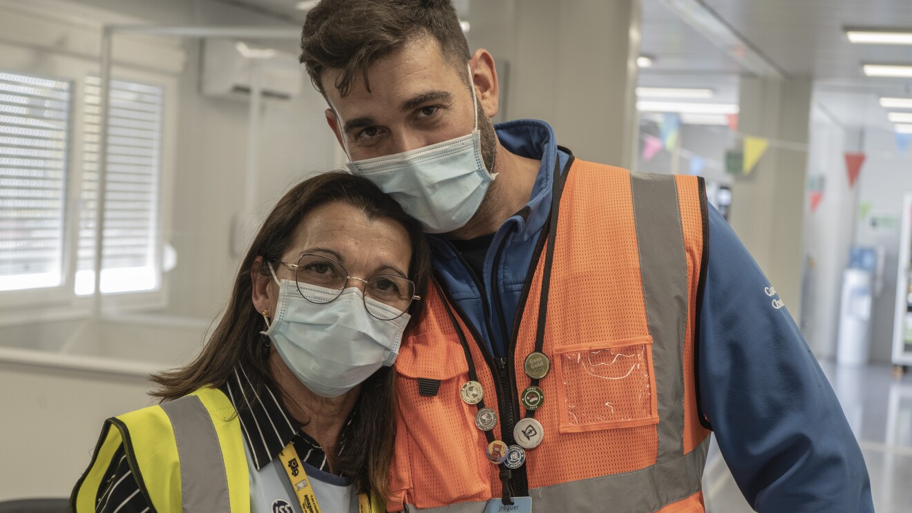 María José Requera y Julio Becerra Requera. Situados en las oficinas de la estación logística de Amazon en Málaga. La madre va con un chaleco amarillo, camisa negra con rayas blancas. Lleva gafas, mascarilla quirúrgica y el pelo suelo por el hombro. Su hijo le pasa la cabeza, lleva mascarilla quirúrgica, el pelo corto, un forro polar azul y un chaleco naranja.