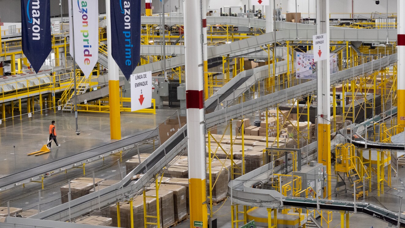Foto con perspectiva de un centro logístico de Amazon. Se puede apreciar solo una personas caminando con un todo semimecánico. El centro tiene las estructuras en amarillo y hay cintas transportadoras de pedidos. Del techo bajan tres carteles al estilo de bandera, en uno pone Amazon, en el otro Amazon Pride y en el tercero Amazon Prime.
