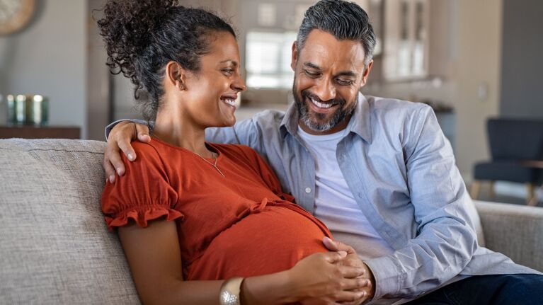 Expectant couple smiling on couch, woman in red top touching belly