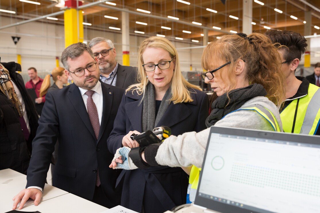 Eine Amazon Mitarbeiterin steht neben der Ministerin, die von weiteren Mitarbeitern und Gästen umringt ist.