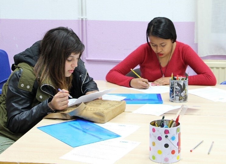 En una mesa blanca aparecen dos chicas estudiando. En la mesa hay hojas, dos carpetas azules y dos vasos con lápices y rotuladores. Una de las chicas lleva el abrigo puesto y lleva puesta una chaqueta negra y verde. Ella está sujetando una mano con un bolígrafo y en la otra tiene el papel. Está leyendo. El papel está sobre su bolso, que está encima de la mesa. Esta chica tiene el pelo oscuro y largo. La otra chica tiene un jersey rojo y está con un lápiz escribiendo en un papel.