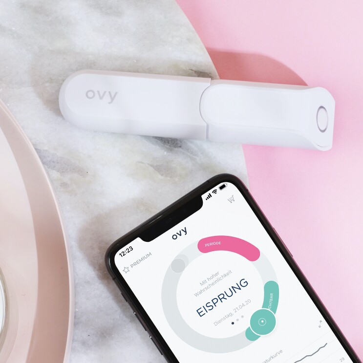 Ein Handy und ein Thermometer liegen auf einer rosanen Unterlage.