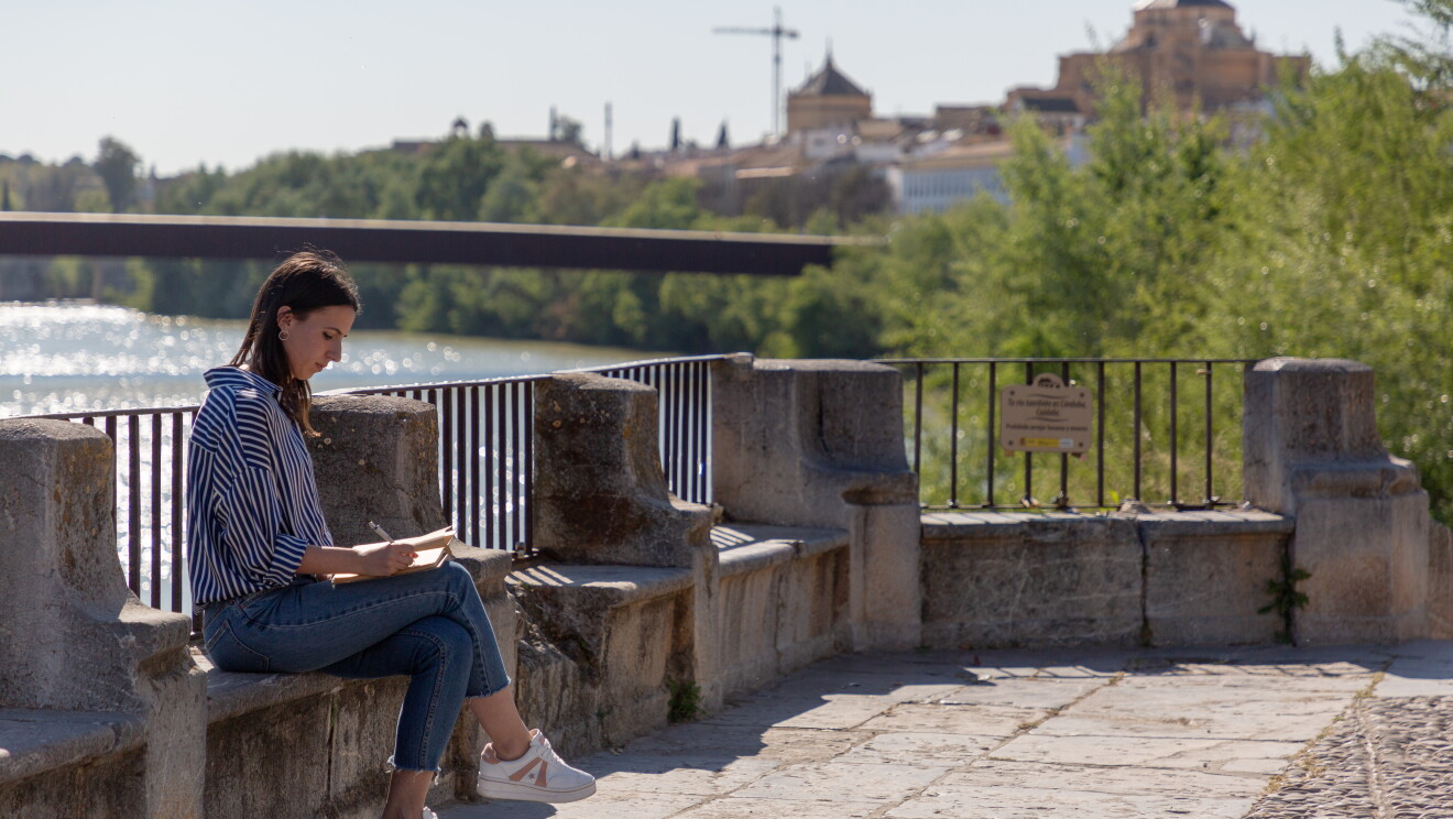 Sonia Llamas es de Córdoba y se encuentra sentada en un puente del Guadalquivir con una libreta en la mano e ilustrando alguna de sus creaciones que pronto venderá en Amazon. Con el pelo largo, una camisa a rayas blanca y azul y unas deportivas blanca. De fondo el río Guadalquivir y parte de la silueta de la ciudad de Córdoba. 