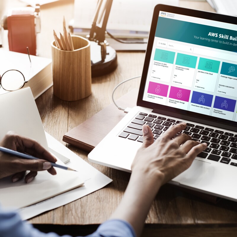 Una mujer racializada en primer plano desenfocada delante de un ordenador portátil. En el ordenador portátil está abierta una pantalla de AWS skill builder. En la mesa de madera hay unas gafas, unos lápices también de madera, una libreta y el pie de una lámpara de mesa.