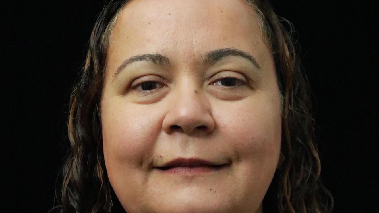 Dilek Hakkani-Tür, a senior principal scientist in the Alexa AI group smiles at the camera. She wears a gray button down shirt and her brown hair is pulled back in a low pony tail.
