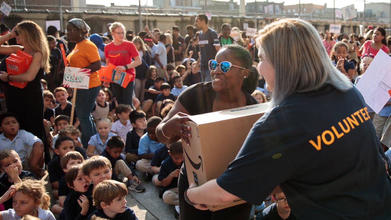 Two adult women carry a cardboard box marked with an Amazon logo. Seated children take up most of the image.