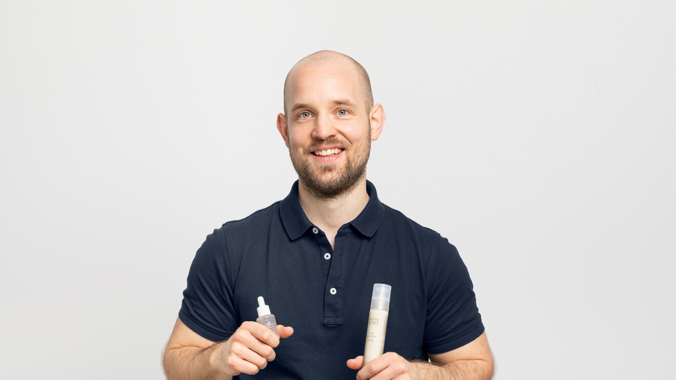 Ein Mann mit Glatze und einem schwarzen Poloshirt lacht in die Kamera. In seinen Händen hält er Produkte von Mellow Noir.