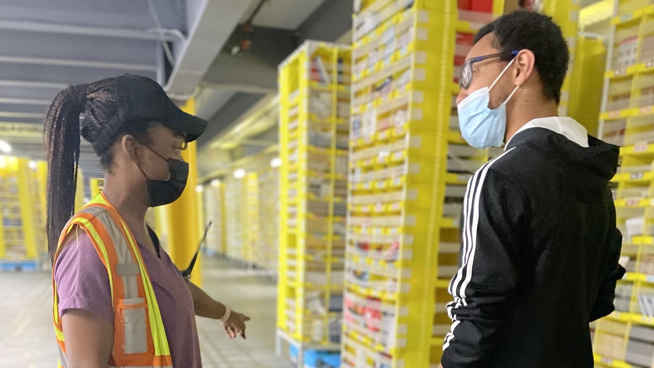 A woman wearing a safety vest in an Amazon fulfillment center (warehouse) speaks to her coworker