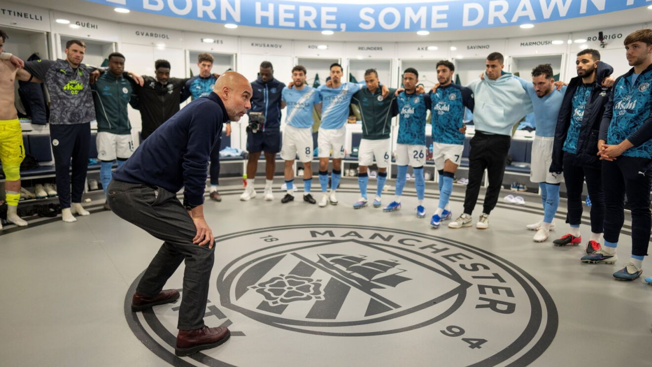 Pep Guardiola stands in the Man City dressing room, giving his players a team talk 