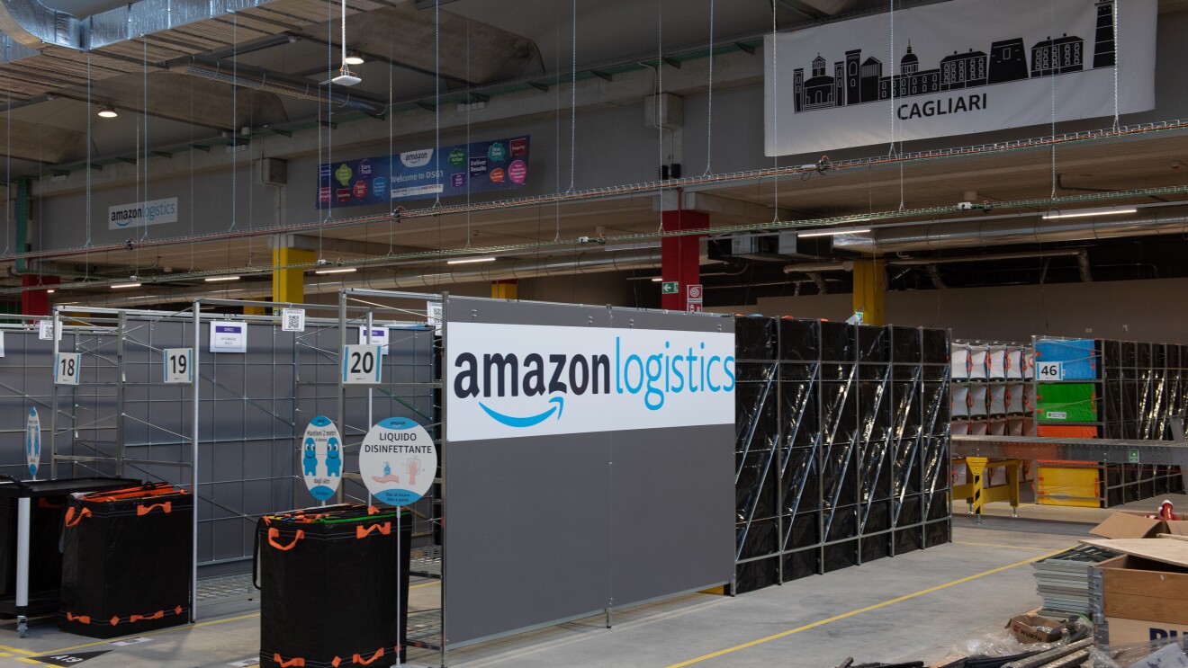 Foto di un'area di un magazzino Amazon. Su un pannello al centro si vede il logo di Amazon Logistics, mentre in fondo uno striscione con il profilo di una città e la scritta Cagliari