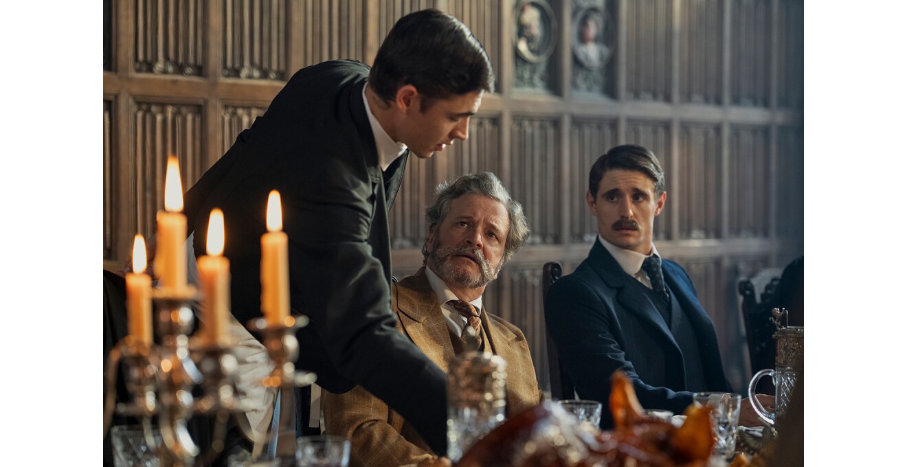 装飾的な羽目板壁の食堂、立って腕を伸ばす男性と、右側に座った2人の男性。座った2人は立っている若者を見ている。