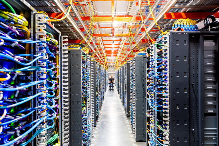 薄暗いデータセンターの屋内の通路。通路の両側にサーバーが多数並び、水色のコードが出ている。