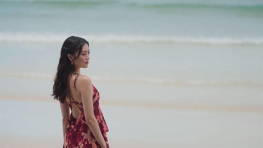 波が打ち寄せる浜辺に立つ女性。紅色の水着とビーチウェアを着用。