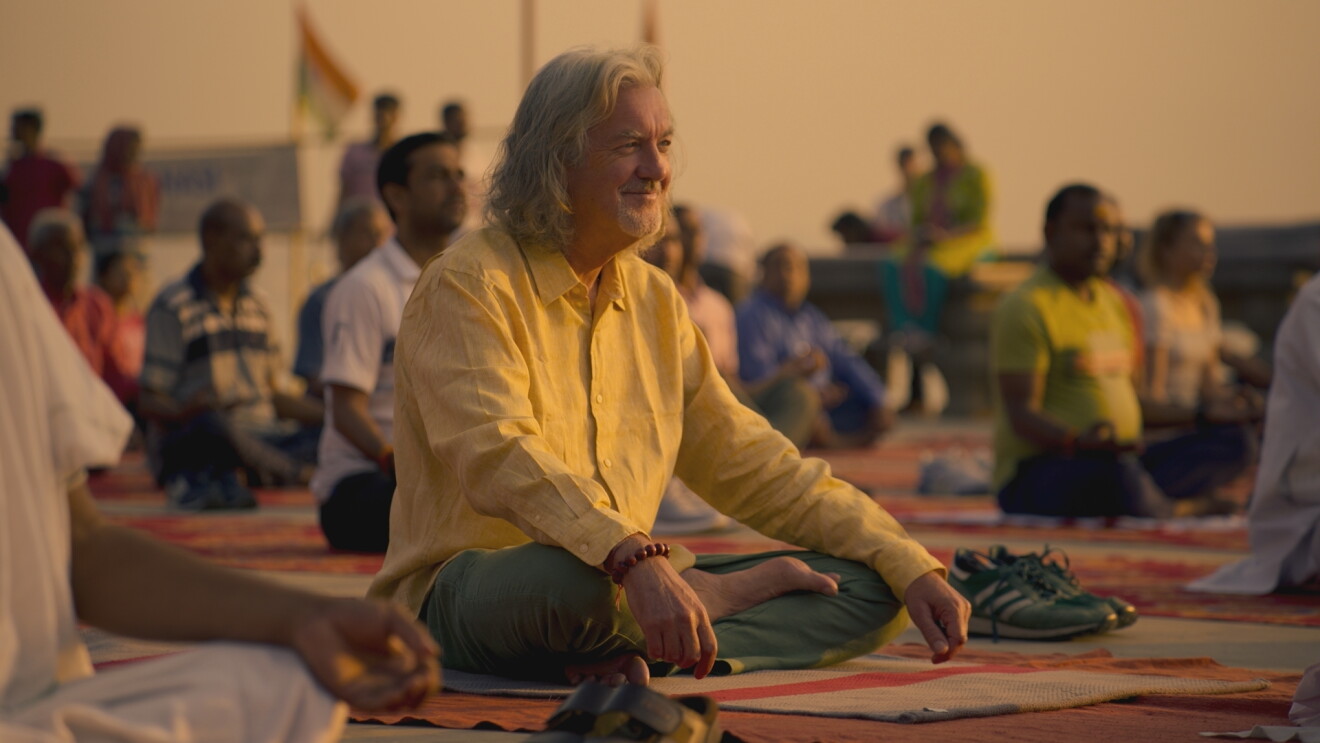 James May sits crossed leg, smiling and meditating with a group in India