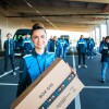 Eine zierliche junge Frau steht im Vordergrund mit einem Amazon Paket, hinter ihr sind 30 Mitarbeiter:innen verteilt auf einem Parkplatz zu sehen. Alle tragen Arbeitskleidung im Amazon Fahrer:innen Branding.