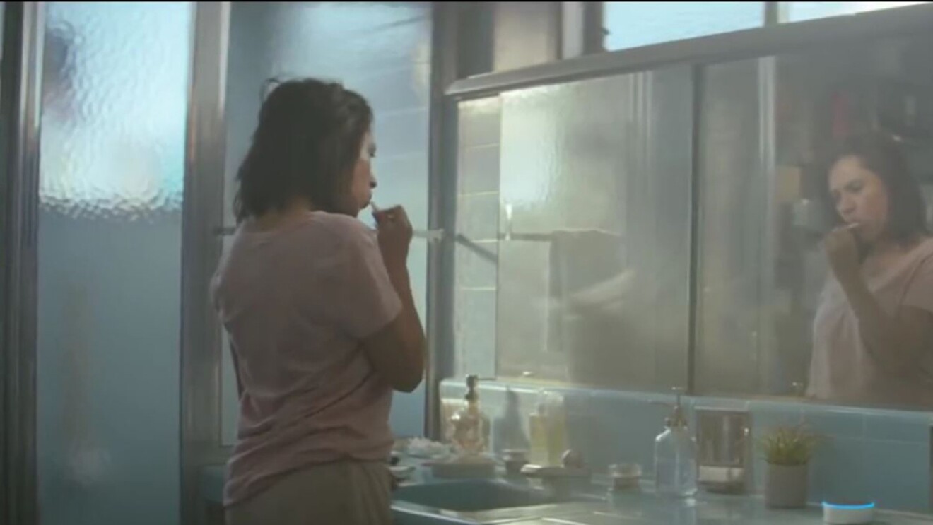 一头黑发,装在一个粉红色的衬衫和运动裤刷她的牙齿在镜子亚马逊回声点落在柜台上。计数器是1970年代蓝色瓷砖。