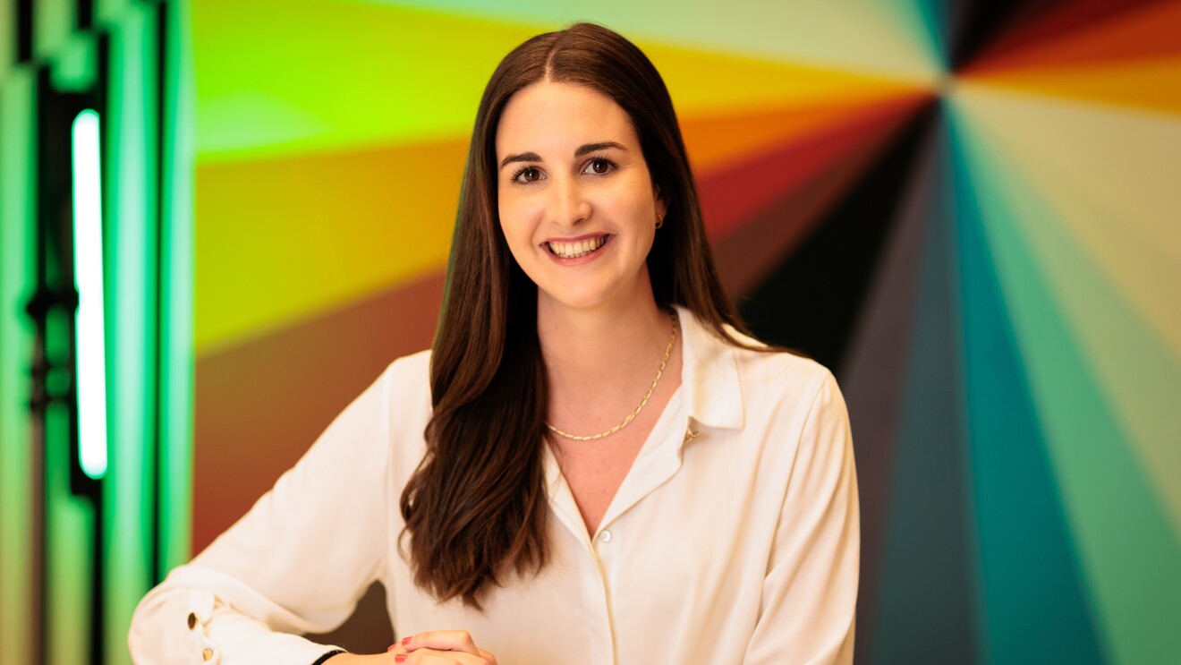 Miriam Domínguez-Adame Aguilera con el pelo liso negro hasta el pecho y una camisa de color blanco. Lleva un collar y un reloj en la mano izquierda. Un fondo de colores con un punto de fuga. Está apoyando las manos en una mesa alta.