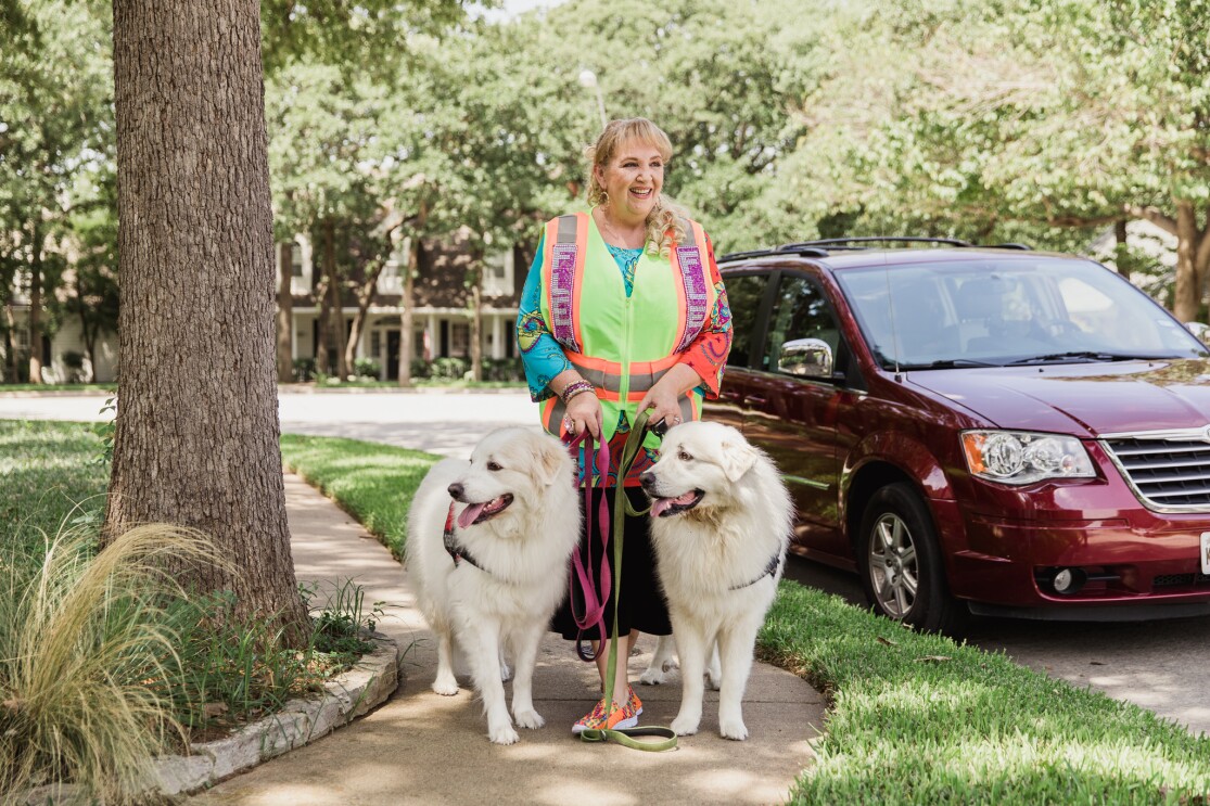 A woman in a bedazzled Amazon Flex vest stands in front of her maroon mini-van. She is smiling at the camera and standing with her two white dogs, who are wearing service animal vests.