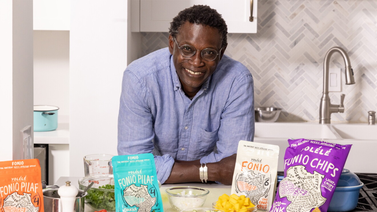 An image of a man smiling for a photo in his kitchen. There are cooking ingredients and four bags of fonio grain products in front of him.