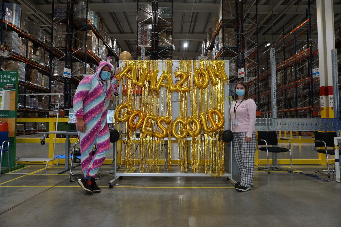 Zwei Mitarbeiter:innen im Pyjama vor einem Schild, auf dem "Amazon goes gold" steht.