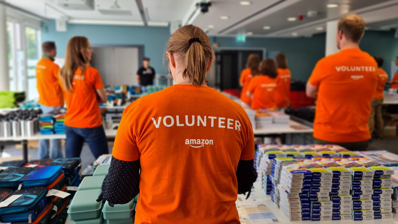 Man sieht Amazon Mitarbeiter:innen mit orangenem Volunteer-T-Shirt von hinten. Sie stehen hinter Tischen mit Schulmaterialien.