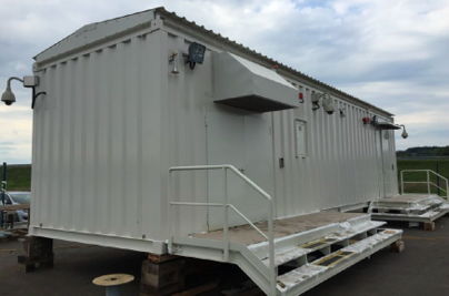 A large white prefabricated modular building (shipping-container style) mounted on steel trailer skids in a paved lot, with a metal staircase and railing leading to a side door. Flat rural/grassy terrain in the background under an overcast sky.
