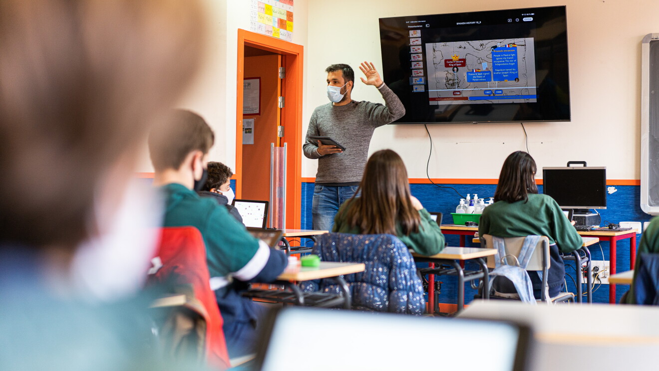Alumnos del IES Josep Tarradellas en una clase con pizarra digital.  es una aula de un centro educativo. Aparacen 5 alumnos de espaldas, sentados en sus pupitres, vestidos con el uniforme de una sudadera de color verde. En la pared una pizarra digital  y  el profesor delante con una tableta en la mano derecha y la izquierda levantada. El profesor tiene barba y el pelo corto y un jersey de color gris y unos tejanos. 