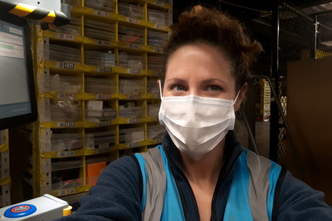 A woman in a safety mask stands with shelving behind her.