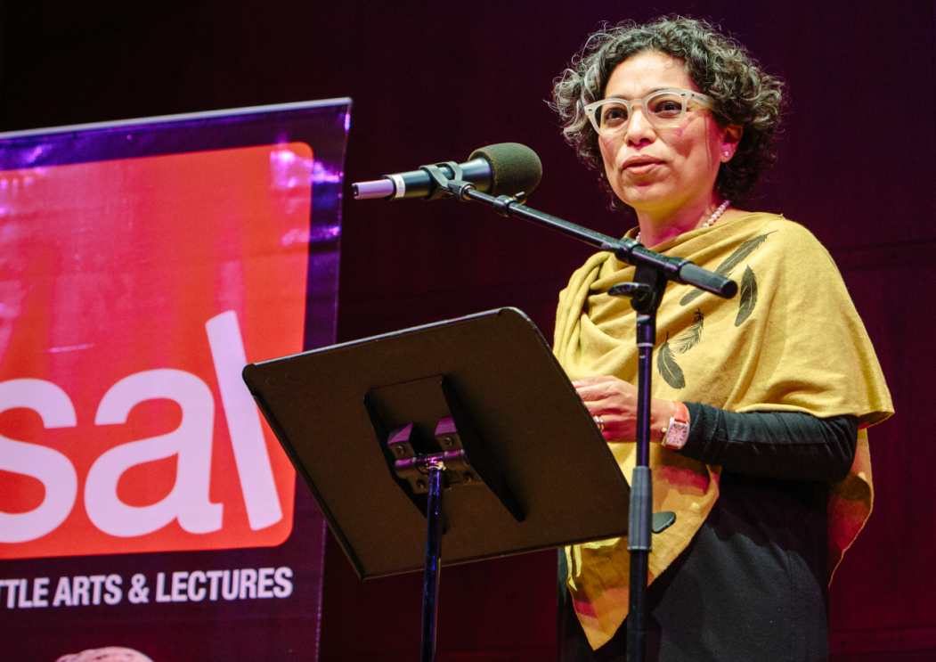 A woman standing at a lecture, in front of a portable podium and microphone. She appears to be speaking to a group, on a stage. In the background, Seattle Arts & Lectures logo is visible.