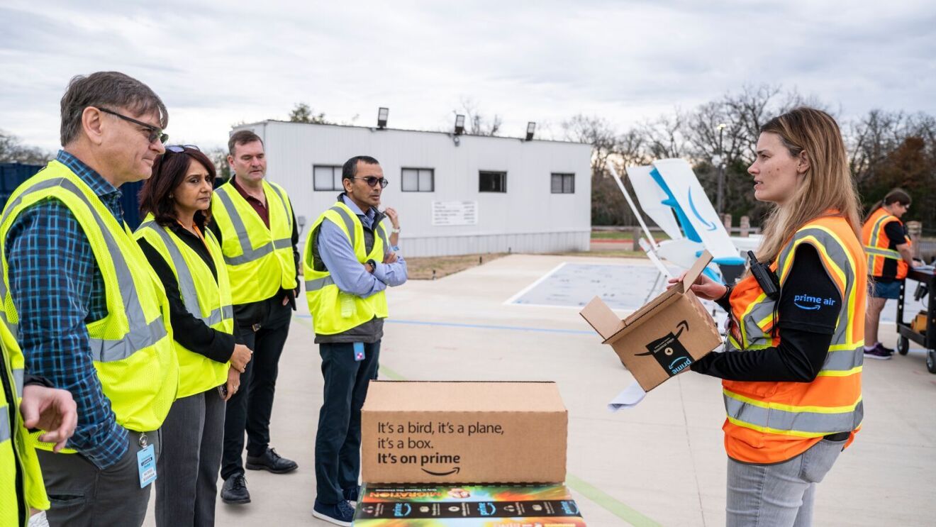 An image of a drone facility employee showing a tour group the custom box design for drone deliveries. They are standing on the landing and launch pad for the drones in Texas.