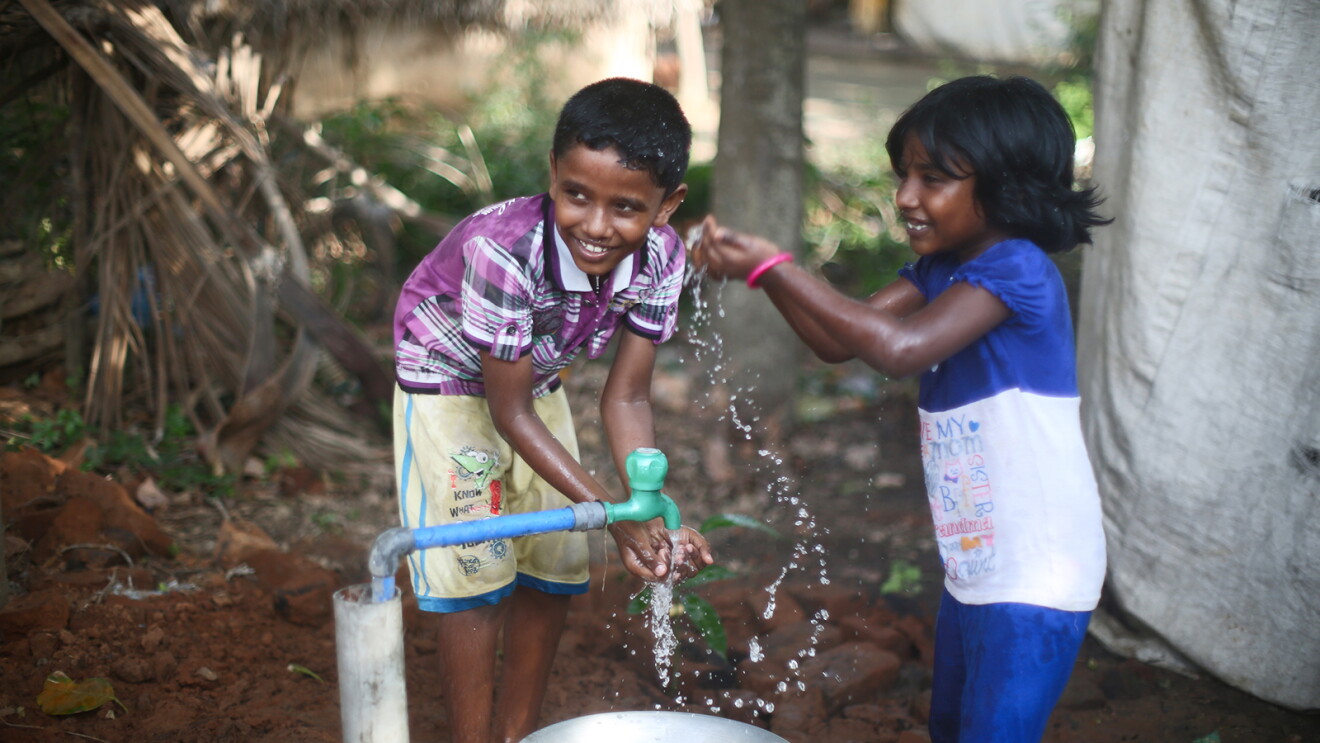 Two children joyfully playing with water from a hand pump in a rural setting