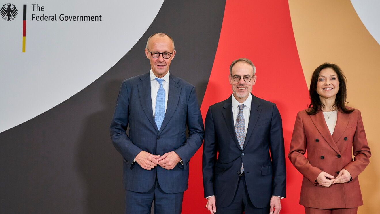Three people stand smiling in front of a backdrop featuring the German Federal Government logo and the black, red and gold colours of the German flag. Two men in dark suits stand beside a woman in a rust-coloured suit.