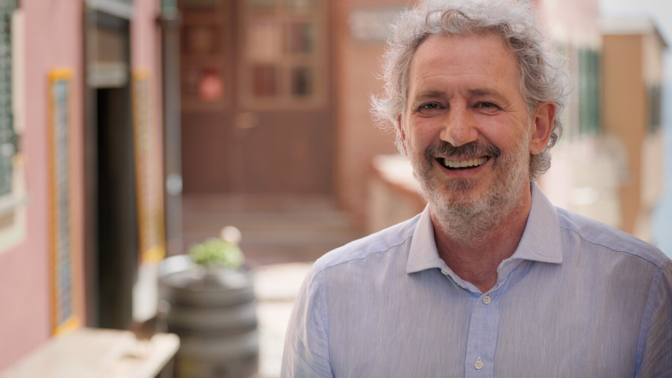 Se ve el centro de un pueblo de Italia, con las fachas de color rosa, un cartel negro con letras blancas que se ve borroso. En primer plano de la foto vemos a Sergio Panizza, de la fábrica Pesto Rossi, de salsa pesto. Sergio tiene el pelo y la barba y el bigote blanco. Está mirando a cámara sonriendo y lleva una camisa de lino azul. 
