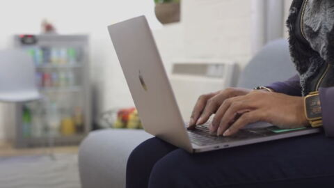 A woman types on the keyboard of a laptop computer while sitting on a sofa. 