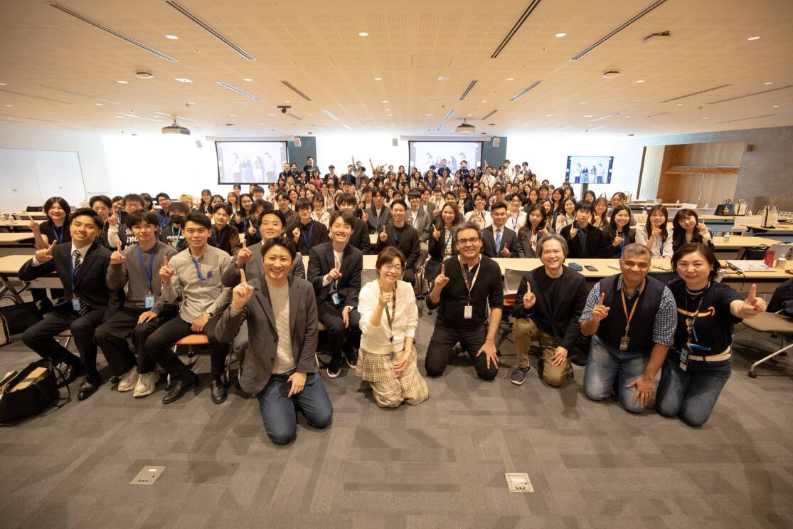 たくさんの人々が広い部屋で集合し、指で「1」のサインをしている