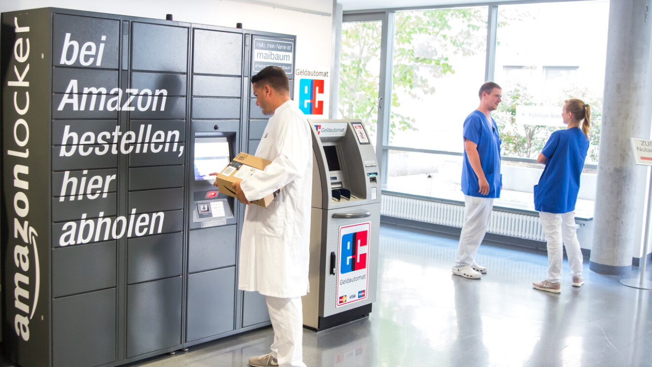 Ein grauer Amazon Locker mit zahlreichen Paketfächern: Am rechten oberen Eck ist zu lesen: "Hallo, mein Name ist maibaum". Ein Arzt mit weißer Hose und weißem Medizinermantel steht vor dem Locker und blickt auf das Eingabedisplay. In der Hand hält er ein Amazon Paket. Rechts neben ihm steht Pflegepersonal: Ein Mann und eine Frau, die sich unterhalten. Beide tragen blaue Arbeitskittel und weiße Hosen.