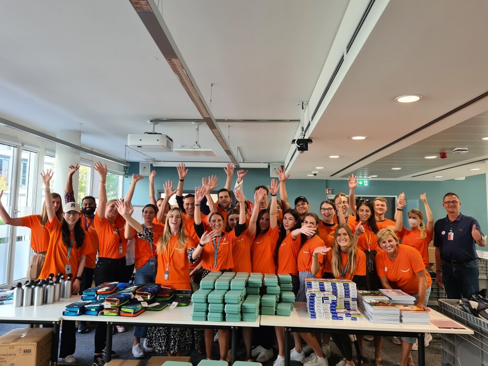 Amazon Mitarbeiter:innen, die sich im Rahmen der "Back to School"- Aktion sozial engagieren. Gruppenbild vor einem Packtisch. Alle Mitarbeiter:innen halten die Hände hoch.