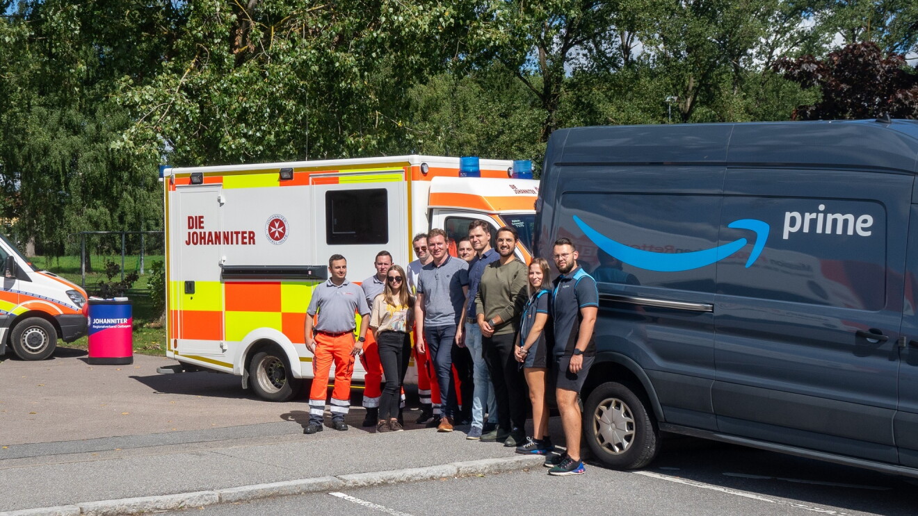 Amazon Lieferfahrerinnen und Lieferfahrer vor einem Amazon Prime Lieferfahrzeug zusammen mit Mitarbeitenden der Johanniter-Unfall-Hilfe vor einem Rettungwagen.
