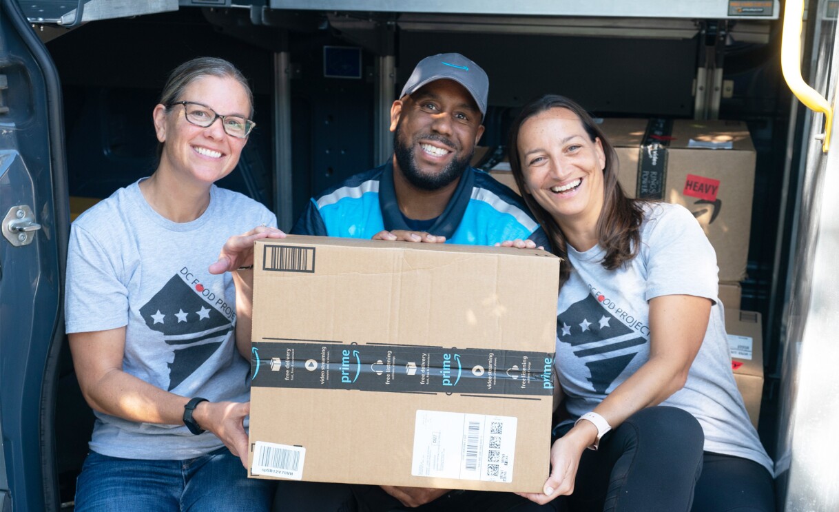 An image of three people sitting on the back of an Amazon Delivery van holding an Amazon delivery box.